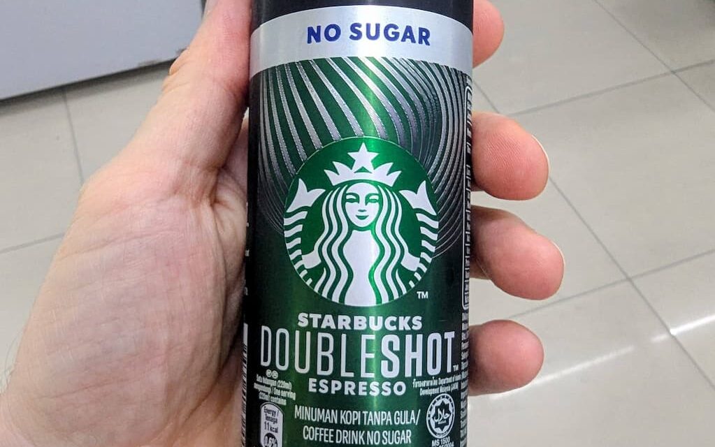 A hand holding a black and silver slim aluminum can of Starbucks Doubleshot Espresso Americano No Sugar inside a 7-Eleven store in Thailand.