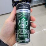 Can You Get a Thailand eSIM at 7-Eleven? (What You Need to Know Before You Walk In) A hand holding a black and silver slim aluminum can of Starbucks Doubleshot Espresso Americano No Sugar inside a 7-Eleven store in Thailand.
