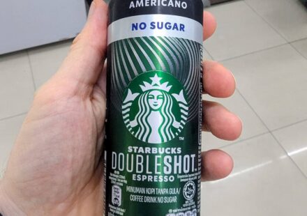 A hand holding a black and silver slim aluminum can of Starbucks Doubleshot Espresso Americano No Sugar inside a 7-Eleven store in Thailand.
