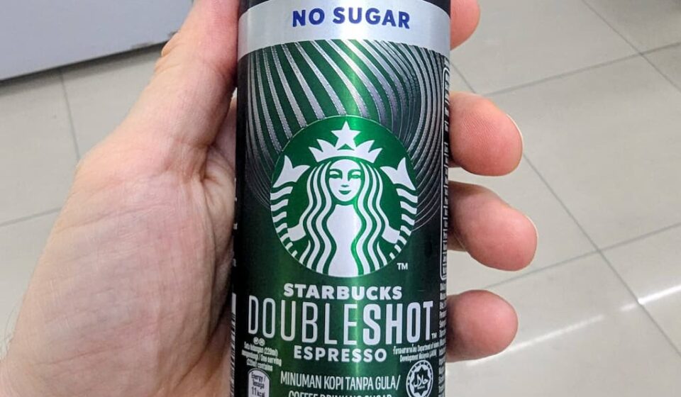 Starbucks Doubleshot Americano No Sugar: Premium Brand Hype Over Mediocre Canned Coffee A hand holding a black and silver slim aluminum can of Starbucks Doubleshot Espresso Americano No Sugar inside a 7-Eleven store in Thailand.