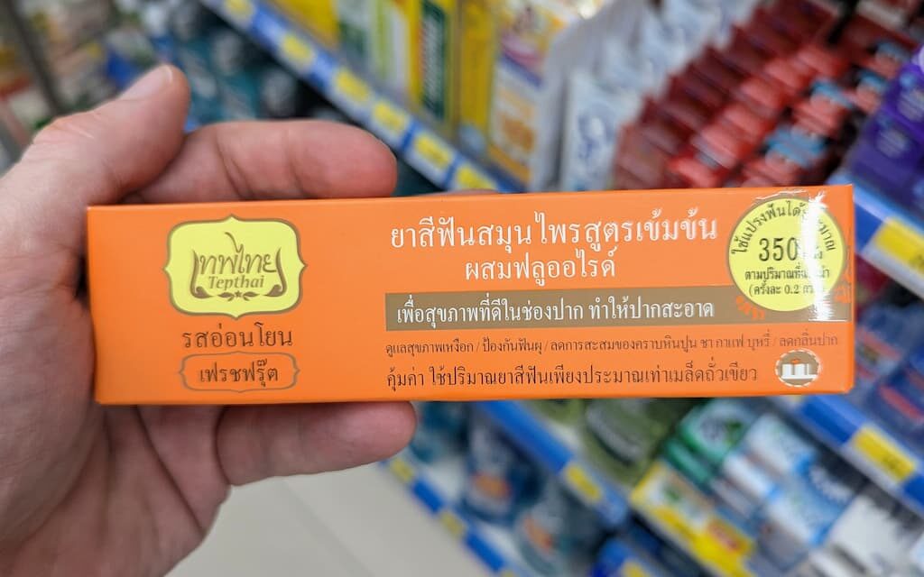 A hand holding an orange box of Tepthai Concentrated Herbal Toothpaste Fresh Fruit with Fluoride inside a 7-Eleven in Thailand.