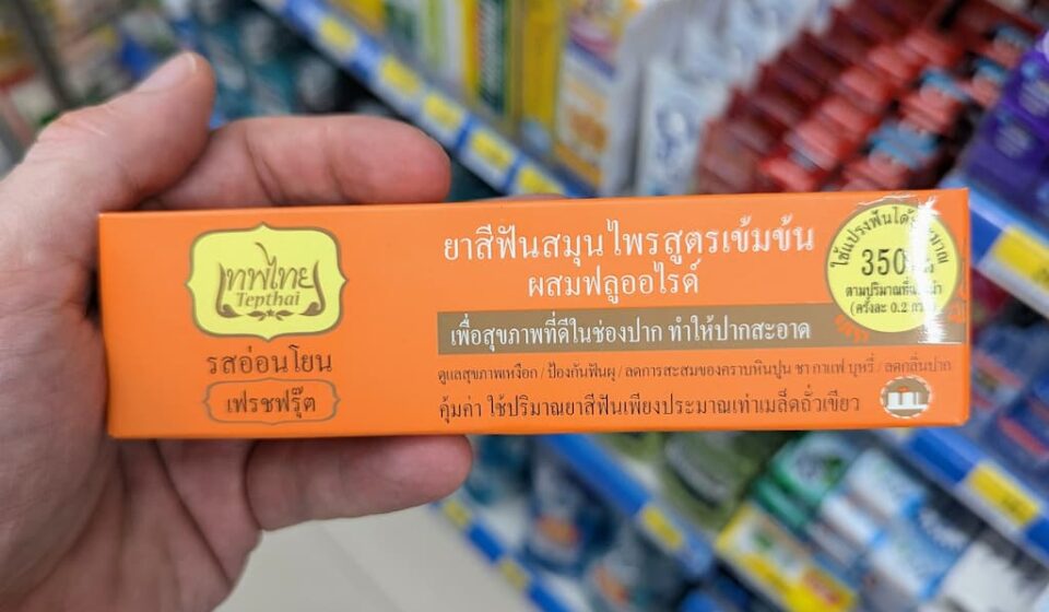 Tepthai Concentrated Herbal Toothpaste Fresh Fruit: High-Yield Oral Care with a Polarizing Fruity Twist A hand holding an orange box of Tepthai Concentrated Herbal Toothpaste Fresh Fruit with Fluoride inside a 7-Eleven in Thailand.