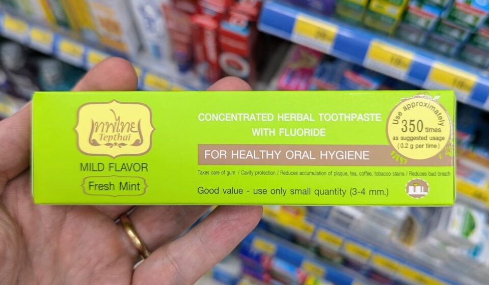 A hand holding a green box of Tepthai Concentrated Herbal Toothpaste Fresh Mint with Fluoride inside a 7-Eleven in Thailand.