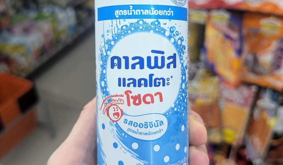 Calpis Lacto Soda Original (Low Sugar): Sweet, Bubbly Japanese Cultured Milk Refreshment A hand holding a slim white and blue polka-dot can of Calpis Lacto Soda Original Low Sugar inside a 7-Eleven store in Thailand.