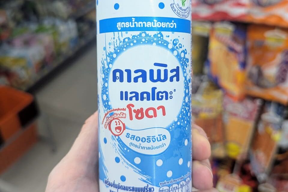 A hand holding a slim white and blue polka-dot can of Calpis Lacto Soda Original Low Sugar inside a 7-Eleven store in Thailand.