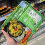 CUP TO GO Papaya Salad with Salted Egg: A Convenience Store Take on Thailand’s Iconic Som Tam A hand holding a green tray of Chef Cares Green Curry with Chicken Breast and Jasmine Rice inside a 7-Eleven store in Thailand.