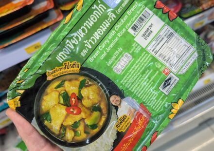 A hand holding a green tray of Chef Cares Green Curry with Chicken Breast and Jasmine Rice inside a 7-Eleven store in Thailand.