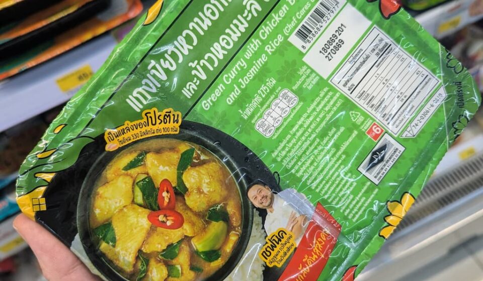 Chef Cares Green Curry with Chicken Breast & Jasmine Rice: A Chef-Crafted Gourmet Lunch at 7-Eleven A hand holding a green tray of Chef Cares Green Curry with Chicken Breast and Jasmine Rice inside a 7-Eleven store in Thailand.