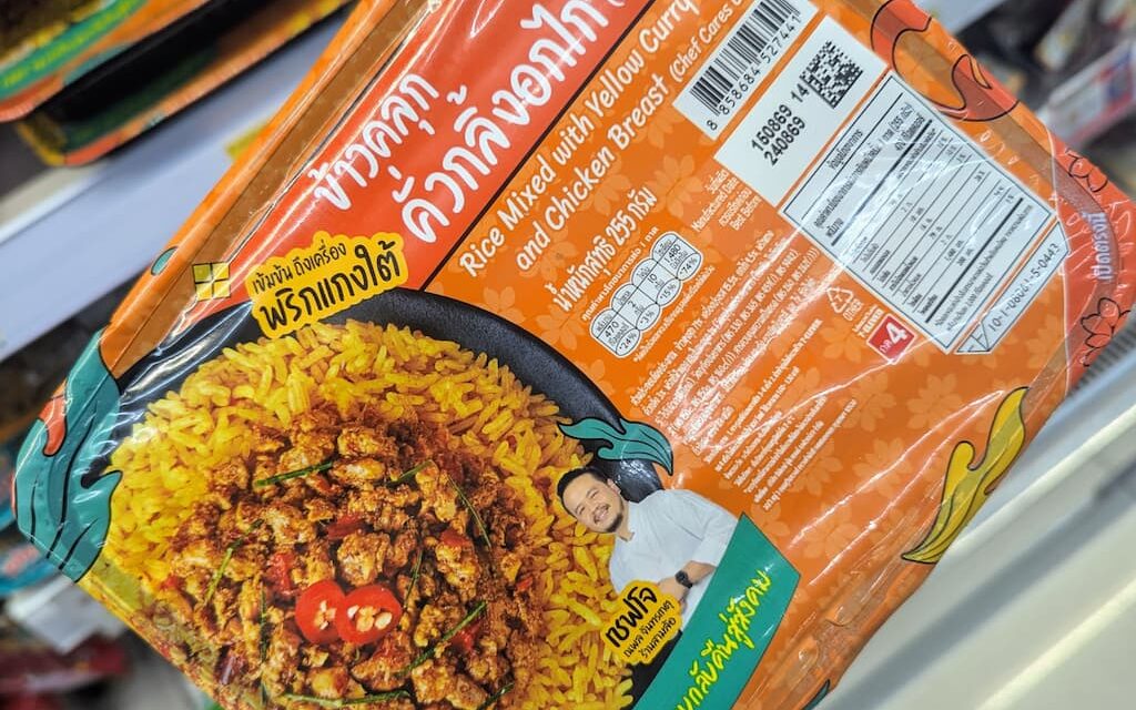 A hand holding an orange tray of Chef Cares Rice Mixed with Yellow Curry Paste and Chicken Breast inside a 7-Eleven store in Thailand.