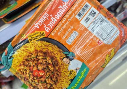 A hand holding an orange tray of Chef Cares Rice Mixed with Yellow Curry Paste and Chicken Breast inside a 7-Eleven store in Thailand.
