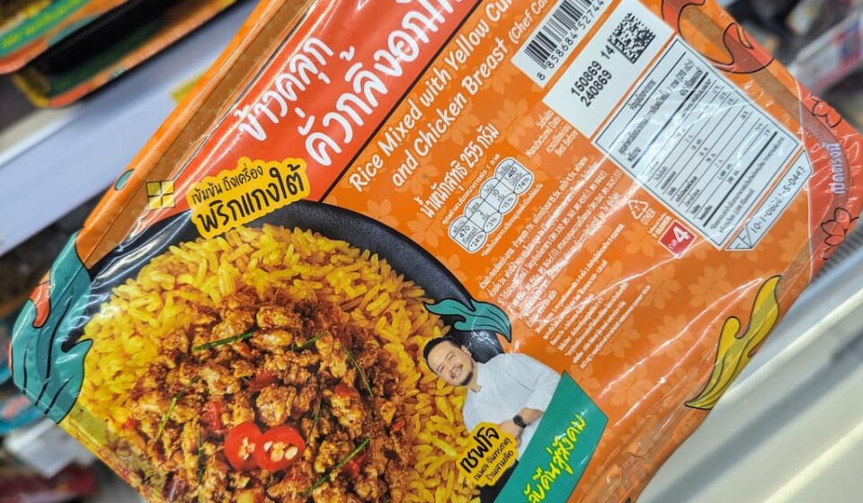 A hand holding an orange tray of Chef Cares Rice Mixed with Yellow Curry Paste and Chicken Breast inside a 7-Eleven store in Thailand.