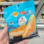 7-Eleven Thailand Monchhichi Stamp Promotion 2026: Complete Premium Collectibles & Expat Redemption Guide A hand holding a cyan-blue package of CP x Ezygo Fish Pocket inside a 7-Eleven store in Thailand.