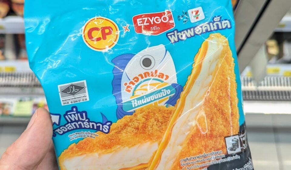 CP x Ezygo Fish Pocket: A Breadless, Tartar-Packed Crispy Fish Treat at 7-Eleven A hand holding a cyan-blue package of CP x Ezygo Fish Pocket inside a 7-Eleven store in Thailand.