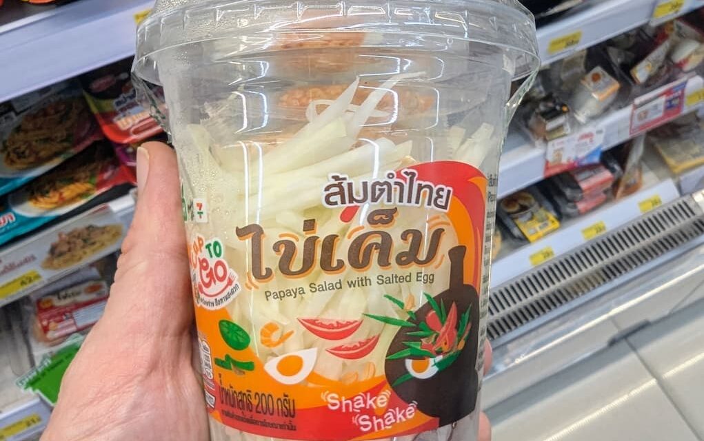A hand holding a plastic cup of CUP TO GO Papaya Salad with Salted Egg inside a 7-Eleven in Thailand.