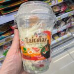 Vitamilk Double Black Sesame & Black Rice: Nutrient-Dense Plant Milk with a Satisfying Roasted Flavor A hand holding a plastic cup of CUP TO GO Papaya Salad with Salted Egg inside a 7-Eleven in Thailand.