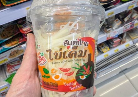 A hand holding a plastic cup of CUP TO GO Papaya Salad with Salted Egg inside a 7-Eleven in Thailand.