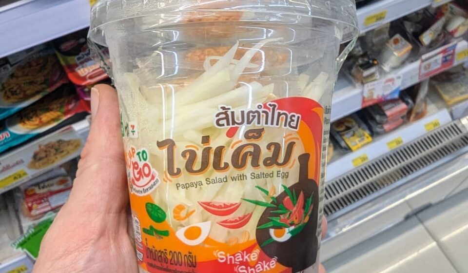 CUP TO GO Papaya Salad with Salted Egg: A Convenience Store Take on Thailand’s Iconic Som Tam A hand holding a plastic cup of CUP TO GO Papaya Salad with Salted Egg inside a 7-Eleven in Thailand.