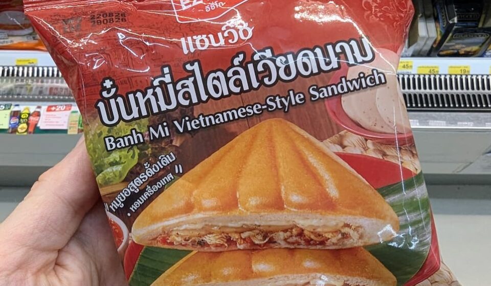 Ezygo Banh Mi Vietnamese-Style Sandwich: A Crunchy Asian Fusion Toastie That Hits the Spot A hand holding a red package of Ezygo Banh Mi Vietnamese-Style Sandwich in front of a 7-Eleven display in Thailand.