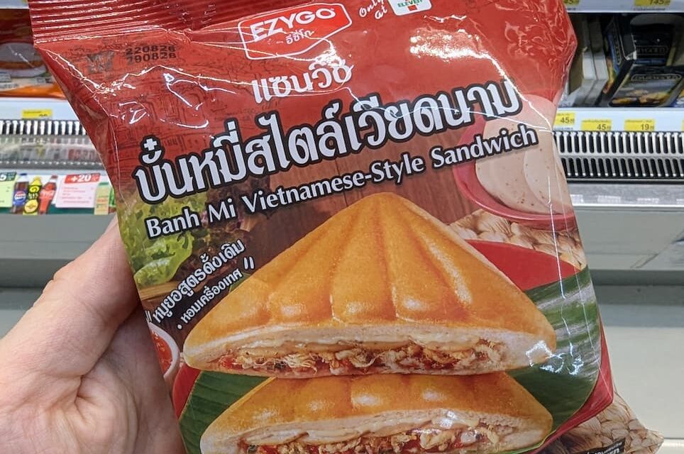 A hand holding a red package of Ezygo Banh Mi Vietnamese-Style Sandwich in front of a 7-Eleven display in Thailand.