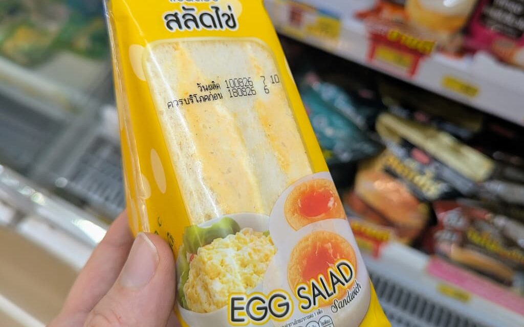 A hand holding a yellow triangle pouch of Ezygo Egg Salad Sandwich in front of a 7-Eleven refrigerator in Thailand.