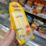 A hand holding a yellow triangle pouch of Ezygo Egg Salad Sandwich in front of a 7-Eleven refrigerator in Thailand.