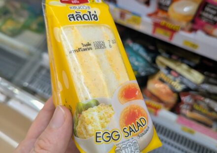 A hand holding a yellow triangle pouch of Ezygo Egg Salad Sandwich in front of a 7-Eleven refrigerator in Thailand.