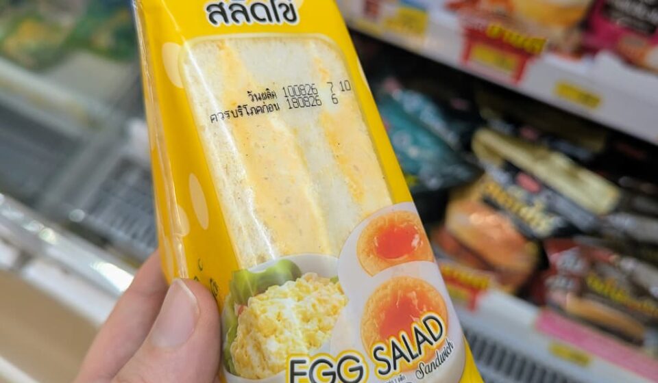 Ezygo Egg Salad Sandwich: Moist, Creamy, and Unbeatable Value for Anytime Snacking A hand holding a yellow triangle pouch of Ezygo Egg Salad Sandwich in front of a 7-Eleven refrigerator in Thailand.
