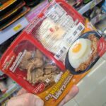 Calpis Lacto Soda Original (Low Sugar): Sweet, Bubbly Japanese Cultured Milk Refreshment A hand holding a red two-compartment tray of Ezygo Garlic Pork and Fried Egg with Rice inside a 7-Eleven store in Thailand.