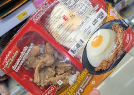 A hand holding a red two-compartment tray of Ezygo Garlic Pork and Fried Egg with Rice inside a 7-Eleven store in Thailand.