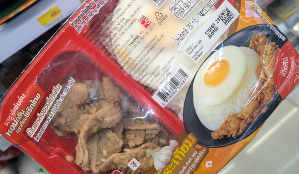 Ezygo Garlic Pork and Fried Egg with Rice: Quick Convenience Spoiled by an Unnatural Fried Egg A hand holding a red two-compartment tray of Ezygo Garlic Pork and Fried Egg with Rice inside a 7-Eleven store in Thailand.