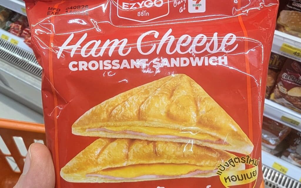 A hand holding a red package of Ezygo Ham Cheese Croissant Sandwich inside a 7-Eleven in Thailand.