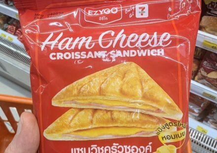 A hand holding a red package of Ezygo Ham Cheese Croissant Sandwich inside a 7-Eleven in Thailand.