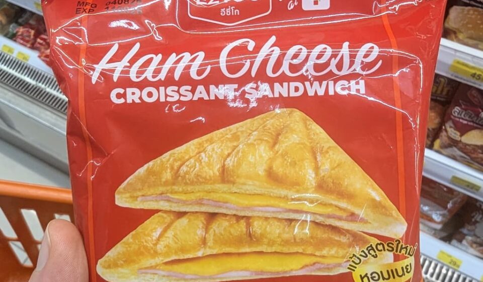 Ezygo Ham Cheese Croissant Sandwich: A Reliable 7-Eleven Classic That Never Fails A hand holding a red package of Ezygo Ham Cheese Croissant Sandwich inside a 7-Eleven in Thailand.