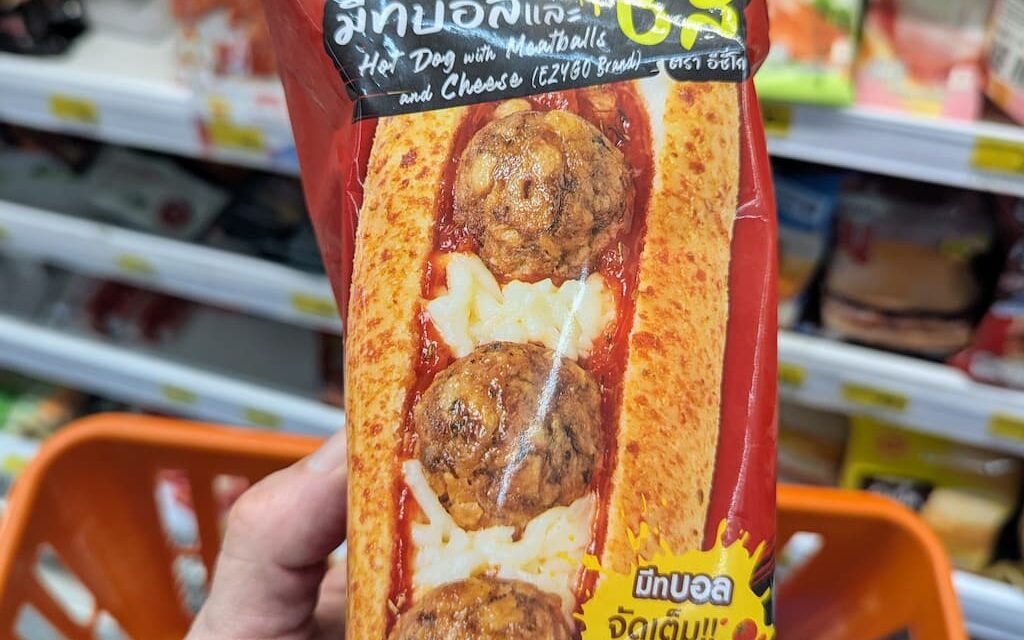 A hand holding a red package of Ezygo Hot Dog with Meatballs and Cheese inside a 7-Eleven store in Thailand.