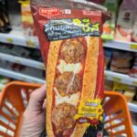 A hand holding a red package of Ezygo Hot Dog with Meatballs and Cheese inside a 7-Eleven store in Thailand.