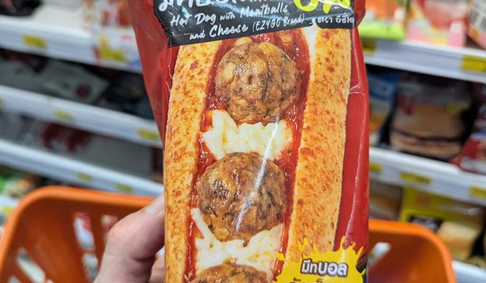 Ezygo Hot Dog with Meatballs and Cheese: A Satisfying Italian-Style Microwave Bit A hand holding a red package of Ezygo Hot Dog with Meatballs and Cheese inside a 7-Eleven store in Thailand.