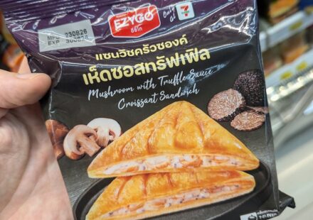 Ezygo Mushroom with Truffle Sauce Croissant Sandwich: A Rich, Gourmet Truffle Indulgence A hand holding a black package of Ezygo Mushroom with Truffle Sauce Croissant Sandwich inside a 7-Eleven store in Thailand.