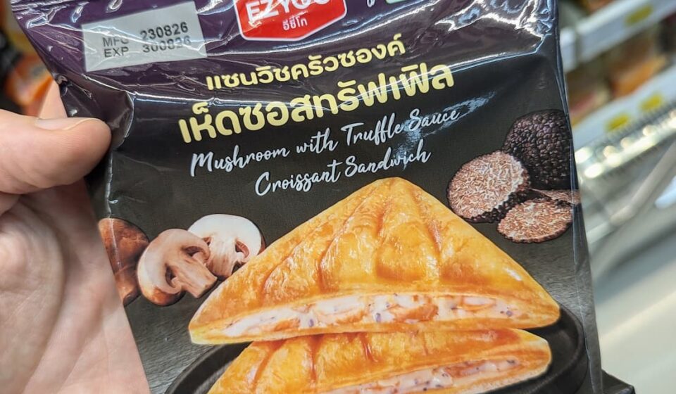 Ezygo Mushroom with Truffle Sauce Croissant Sandwich: A Rich, Gourmet Truffle Indulgence A hand holding a black package of Ezygo Mushroom with Truffle Sauce Croissant Sandwich inside a 7-Eleven store in Thailand.