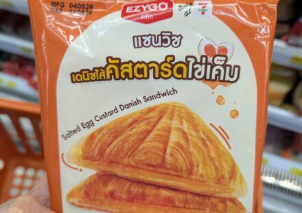 A hand holding an orange package of Ezygo Salted Egg Custard Danish Sandwich inside a 7-Eleven store in Thailand.