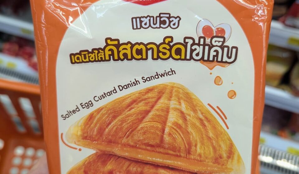 A hand holding an orange package of Ezygo Salted Egg Custard Danish Sandwich inside a 7-Eleven store in Thailand.