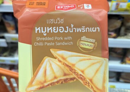 A hand holding a brown and beige package of Ezygo Shredded Pork with Chilli Paste Sandwich inside a 7-Eleven in Thailand.