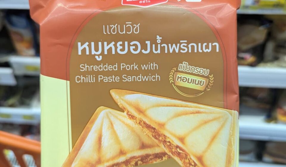A hand holding a brown and beige package of Ezygo Shredded Pork with Chilli Paste Sandwich inside a 7-Eleven in Thailand.