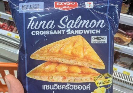 A hand holding a navy-blue package of Ezygo Tuna Salmon Croissant Sandwich in front of a 7-Eleven display in Thailand.