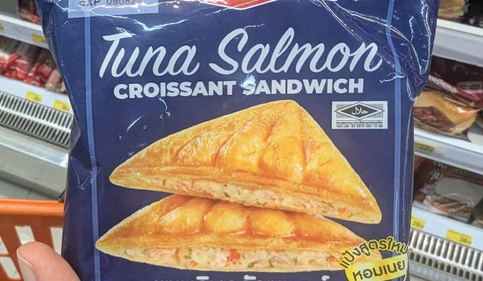 Ezygo Tuna Salmon Croissant Sandwich: Crispy Flaky Exterior with a Moist, Creamy Seafood Center A hand holding a navy-blue package of Ezygo Tuna Salmon Croissant Sandwich in front of a 7-Eleven display in Thailand.