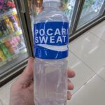 A hand holding a 500ml bottle of Pocari Sweat Ion Supply Drink in front of a 7-Eleven beverage cooler in Thailand.