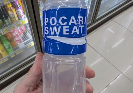 Pocari Sweat Ion Supply Drink: Essential Hydration for Tropical Heat & Post-Illness Recovery A hand holding a 500ml bottle of Pocari Sweat Ion Supply Drink in front of a 7-Eleven beverage cooler in Thailand.
