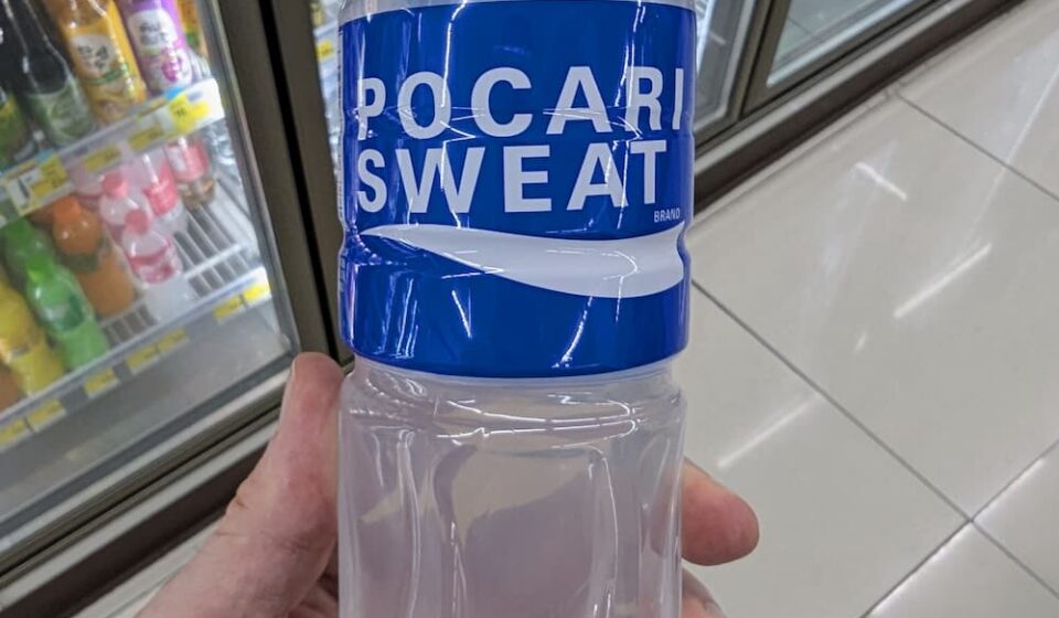 Pocari Sweat Ion Supply Drink: Essential Hydration for Tropical Heat & Post-Illness Recovery A hand holding a 500ml bottle of Pocari Sweat Ion Supply Drink in front of a 7-Eleven beverage cooler in Thailand.