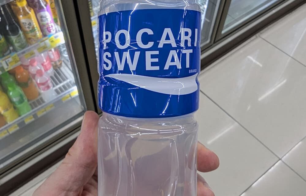 A hand holding a 500ml bottle of Pocari Sweat Ion Supply Drink in front of a 7-Eleven beverage cooler in Thailand.