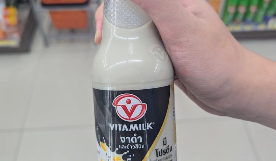 Vitamilk Double Black Sesame & Black Rice: Nutrient-Dense Plant Milk with a Satisfying Roasted Flavor A hand holding a 300ml glass bottle of Vitamilk Double Black Sesame and Black Rice soy milk inside a 7-Eleven in Thailand.