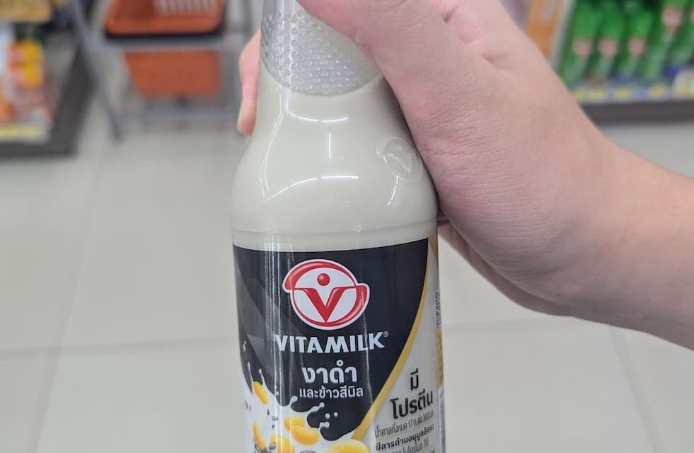 A hand holding a 300ml glass bottle of Vitamilk Double Black Sesame and Black Rice soy milk inside a 7-Eleven in Thailand.